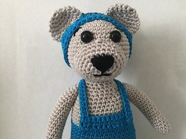 Gehäkelter Amigurumi-Teddy in Beige mit blauem Stirnband, blauer Latzhose und schwarzen Augen
