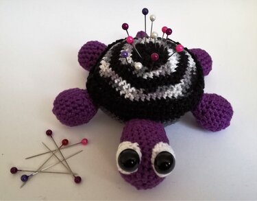 Gehäkeltes Schildkröten-Nadelkissen mit gestreiftem Panzer und lila Gliedmaßen, bestückt mit Stecknadeln auf hellem Hintergrund.