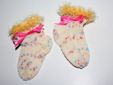 Zwei cremefarbene handgestrickte Babysocken mit bunten Sprenkeln, gelben flauschigen Bündchen und rosa Satinschleifen