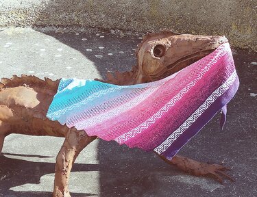 Gestricktes Dreieckstuch in Türkis, Pink und Violett mit Lochmuster, auf rostiger Metallskulptur drapiert.