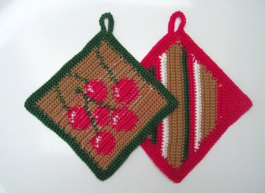 Zwei quadratische Topflappen mit Schlaufen, ein Motiv mit roten Beeren, das andere mit diagonalen Streifen