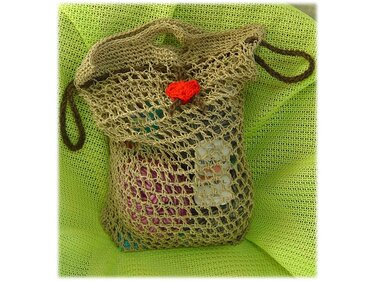 Beigefarbene gehäkelte Netztasche mit braunen Henkeln und rotem Herz, gefüllt mit Garn, auf grünem, strukturiertem Tuch.