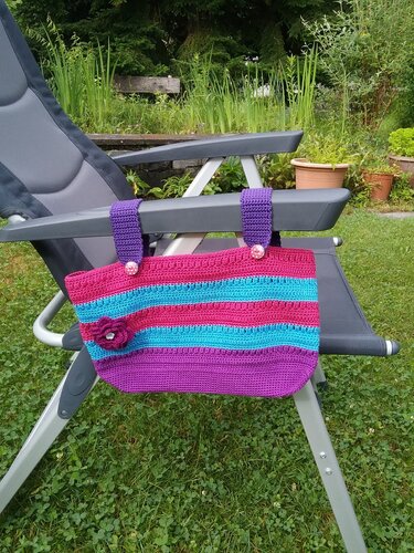 Gehäkeltes Utensilo in Pink, Türkis und Lila mit Schlaufen und Knöpfen, an einer Gartenliege befestigt