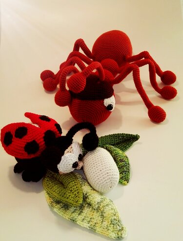 Rote gehäkelte Amigurumi-Spinne, roter Marienkäfer und weißer Kokon auf grünen gehäkelten Blättern