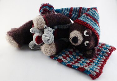 Gehäkelter brauner Bär mit gestreifter Mütze und zwei kleine graue gehäkelte Tiere auf einer bunten Häkeldecke