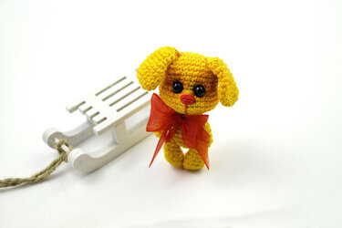 Gelbes gehäkeltes Amigurumi-Hündchen mit roter Schleife neben kleinem weißen Holzschlitten