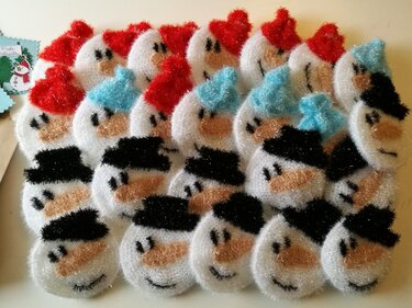 Nahaufnahme von mehreren gehäkelten, runden Schneemann-Gesichtern aus schimmerndem Garn mit verschiedenen Mützenfarben