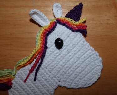 Gehäkelte Einhorn-Applikation mit weißem Körper, schwarzem Auge, violettem Horn und bunter Regenbogenmähne auf Holzhintergrund.