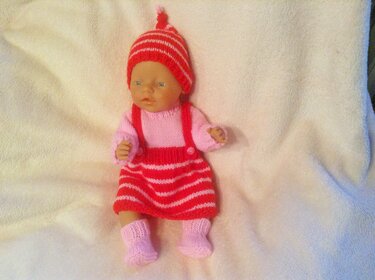 Babypuppe auf heller Decke mit gestricktem rot-rosa gestreiftem Outfit: Zipfelmütze, Latzkleid und Söckchen.
