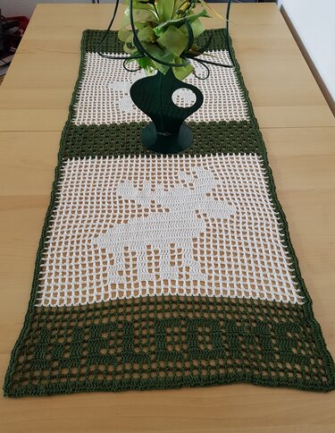 Gehäkelter grüner und weißer Tischläufer im Filetmuster mit Rentiermotiv auf einem Holztisch, dekoriert mit einer grünen Vase.