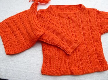 Oranger gestrickter Kinderpullover und passende Mütze mit Bindebändern auf hellem Untergrund