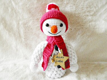 Gehäkelter weißer Schneemann mit roter Mütze und Schal, Holzstern 'Alles Liebe' und Glöckchen auf flauschigem Untergrund.