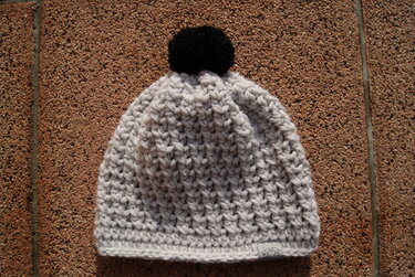 Hellgraue gehäkelte Beanie mit schwarzem Bommel auf Steinboden