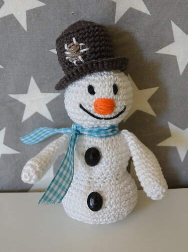Gehäkelter Schneemann mit braunem Hut und blau-weiß kariertem Schal vor Sternenmuster