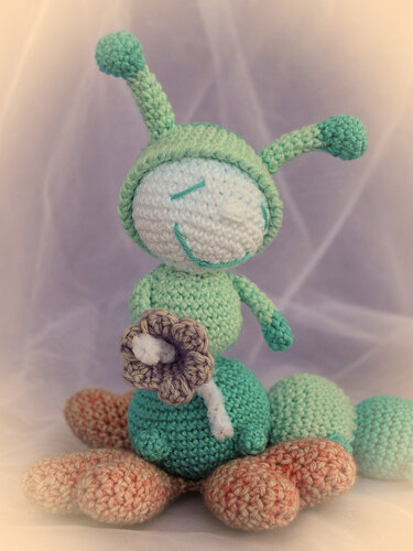 Gehäkelte Amigurumi-Raupe in Türkis und Hellgrün mit lila Blume und pfirsichfarbenen Füßen