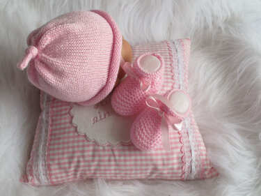 Rosa gestrickte Babymütze und Babyschuhe mit Satinband auf einem karierten Kissen