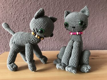 Zwei graue Amigurumi-Katzen aus Garn mit langen Beinen, grünen Sicherheitsaugen und bunten Perlenhalsbändern auf einer Holzfläche vor strukturierter Wand