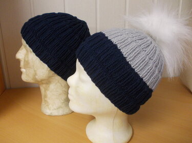 Zwei gestrickte Rippenmützen auf Schaufensterköpfen: eine dunkelblaue, eine blau-graue mit weißem Bommel