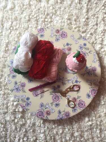 Häkelutensilien auf einer floralen Platte: weiße, rote und rosa Garnknäuel, Häkelnadel, Nähnadel, Schere und gehäkelter Cupcake mit Erdbeere.