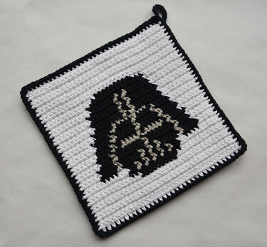 Square crocheted potholder with a bold black helmet motif on a white background, black border and hanging loop.