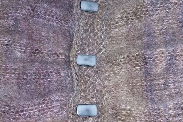 Nahaufnahme eines flauschigen, gestrickten Textils in melierten Lila- und Brauntönen mit drei hellen, rechteckigen Knöpfen.