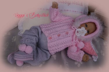 Babypuppe in rosa gestrickter Kapuzenjacke mit Fellrand, grauer Strickhose und rosa Schühchen mit Bommeln