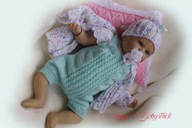 Babypuppe liegt auf Decke und trägt mintfarbenen gestrickten Strampler, bunt gesprenkeltes Jäckchen, passende Mütze und einen Schnuller mit gehäkelter Hülle