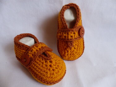 Zwei gehäkelte Babyschuhe in Orange und Braun mit Knopf auf weißem Hintergrund