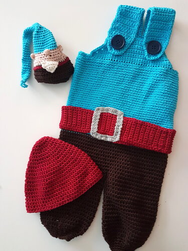 Gehäkeltes Baby-Set: türkise Latzhose mit Knöpfen, roter Bund mit Schnalle, rote Mütze und kleine Zwergenfigur auf hellem Hintergrund.
