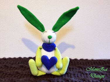 Weiß-grün-blauer gehäkelter Hase in Sitzpose mit Herz auf dem Bauch