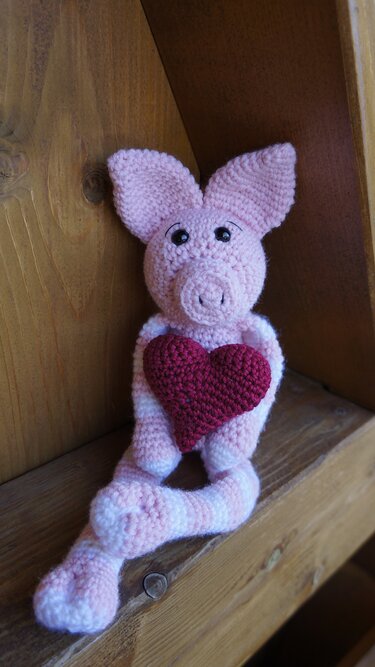 Gehäkeltes rosa Amigurumi‑Schwein mit gestreiften Beinen, das ein rotes Herz hält