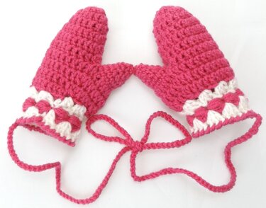 Zwei pinke gehäkelte Babyfäustlinge mit weiß-pinker Zierborte und verbindender Kordel auf hellem Hintergrund.