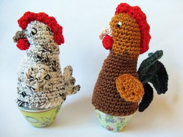 Zwei gehäkelte Eierwärmer in Form eines gesprenkelten Huhns und eines braunen Hahns auf Eierbechern.