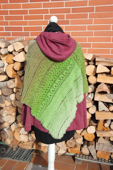 Gehäkelter grüner Poncho mit Farbverlauf und Musterrapport auf einer Schaufensterpuppe vor Holzstapel und Ziegelwand.