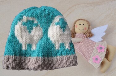 Türkise gestrickte Babymütze mit weißen Schafmotiven und grauem Bündchen neben einer Stoffpuppe