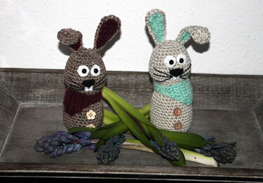 Zwei Amigurumi-Hasen, einer braun und einer hellbeige, mit Schals und Knöpfen auf einem Holztablett neben lila Hyazinthen.