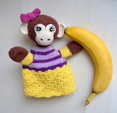 Gehäkelte Affen-Handpuppe mit pinker Schleife, lila-weißem Ringeloberteil und gelbem Rock neben einer Banane