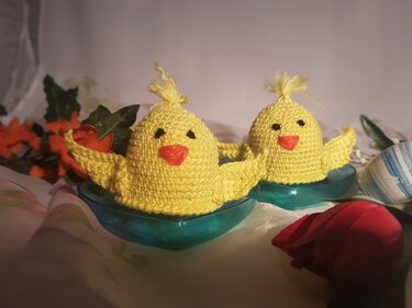Zwei gelbe, gehäkelte Küken-Eierwärmer in türkisfarbenen Schälchen, umgeben von Osterdeko