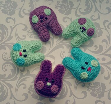 Fünf gehäkelte Hasenfiguren in Lila, Mint und Türkis mit runden Bäckchen und gestickten Gesichtern auf floral gemusterter Unterlage.