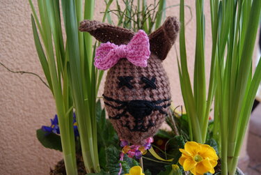 Braunes gehäkeltes Osterhasen‑Ei mit rosa Schleife, gesticktem Gesicht und Perlen zwischen grünen Pflanzen und gelben Blumen.