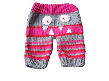 Verspielte gestrickte Babyhose in Rosa und Grau, mit weißen Applikationen und Knopfakzenten, die ein Monstergesicht bilden