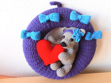 Violetter Häkelring mit hellblauen Schleifen und grauem Amigurumi-Bär, der ein rotes Herz hält