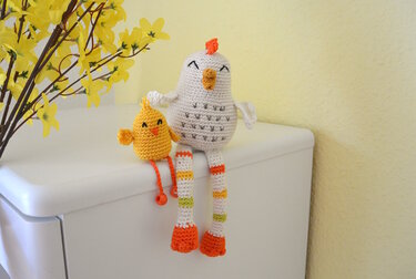 Gehäkeltes Amigurumi-Huhn mit gestreiften Beinen und kleines gelbes Küken auf weißer Kommode