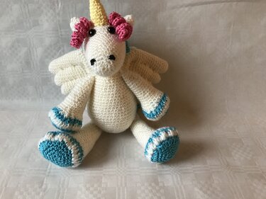 White crocheted unicorn amigurumi with teal-trimmed hooves, yellow horn, pink curly mane and small wings, seated on a light fabric.