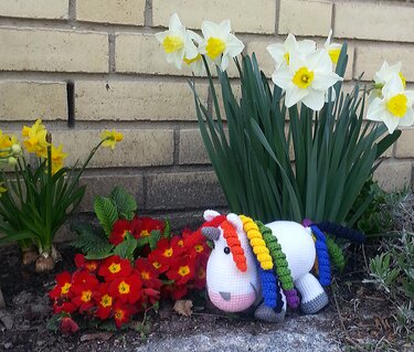 Weißes gehäkeltes Einhorn‑Amigurumi mit Regenbogenmähne im Blumenbeet neben Narzissen