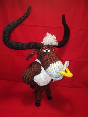 Gehäkeltes braun-weißes Amigurumi-Tier mit langen schwarzen Hörnern und gelbem Nasenring vor rotem Hintergrund