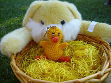 Gehäkelter gelber Küken‑Schlüsselanhänger in einem Weidenkorb mit gelbem Ostergras vor einem großen gelben Plüschhasen.