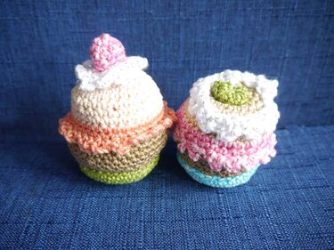 Zwei gehäkelte Cupcakes in Pastellfarben auf blauem Stoff, einer mit pinker Deko, einer mit grünem Detail.