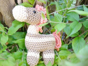 Gehäkeltes Amigurumi-Pferd in hellen Brauntönen mit rosa Mähne und Schweif, platziert zwischen grünen Blättern.