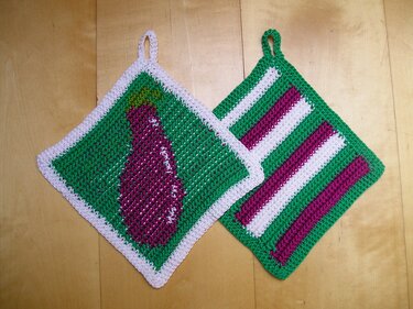 Zwei gehäkelte Topflappen: einer mit lila Aubergine auf grün, der andere gestreift in Grün, Weiß und Lila.